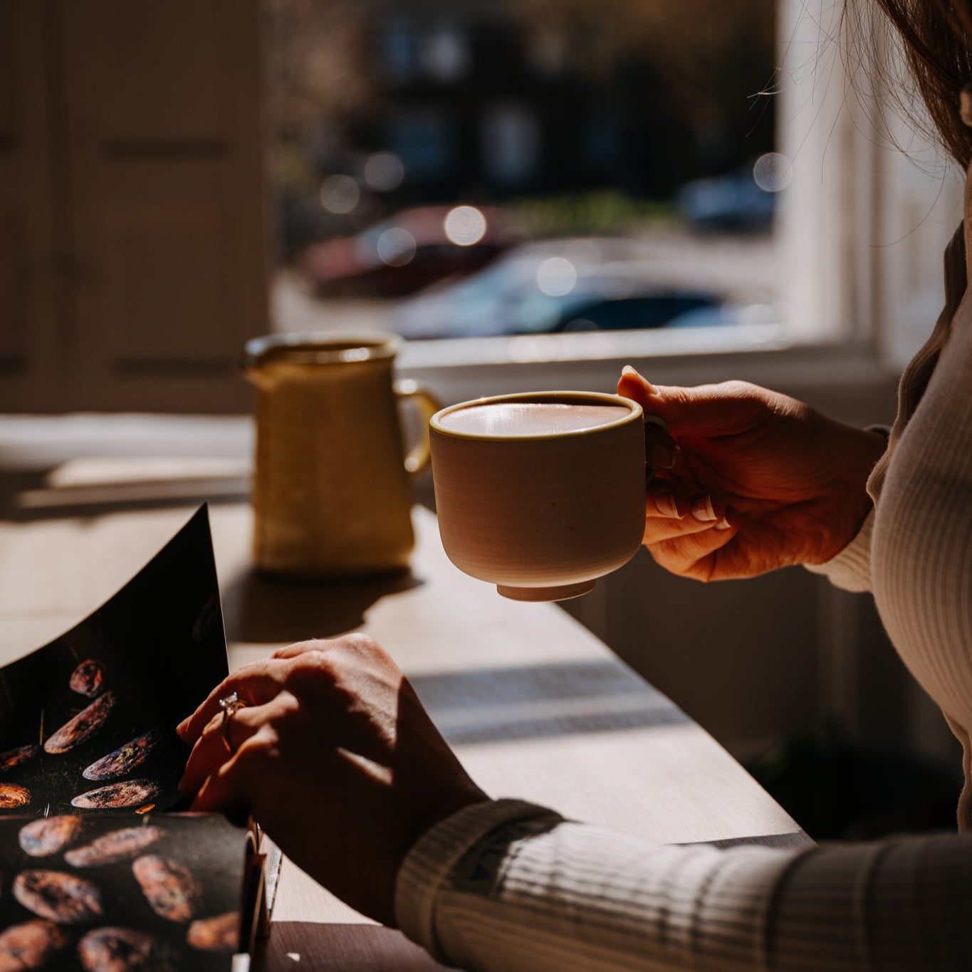Person holding a mug of cacao and looking at a book with cacao bean pictures in a cosy setting.
