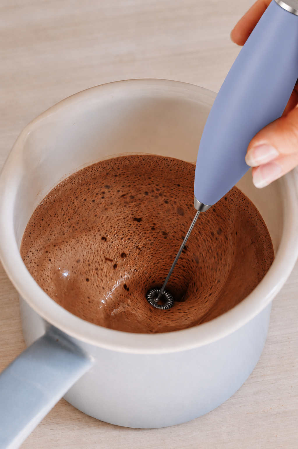 A close-up of a hand holding a light blue handheld electric frother over a small white pot containing frothy brown liquid, likely milk or coffee, on a light wood surface.