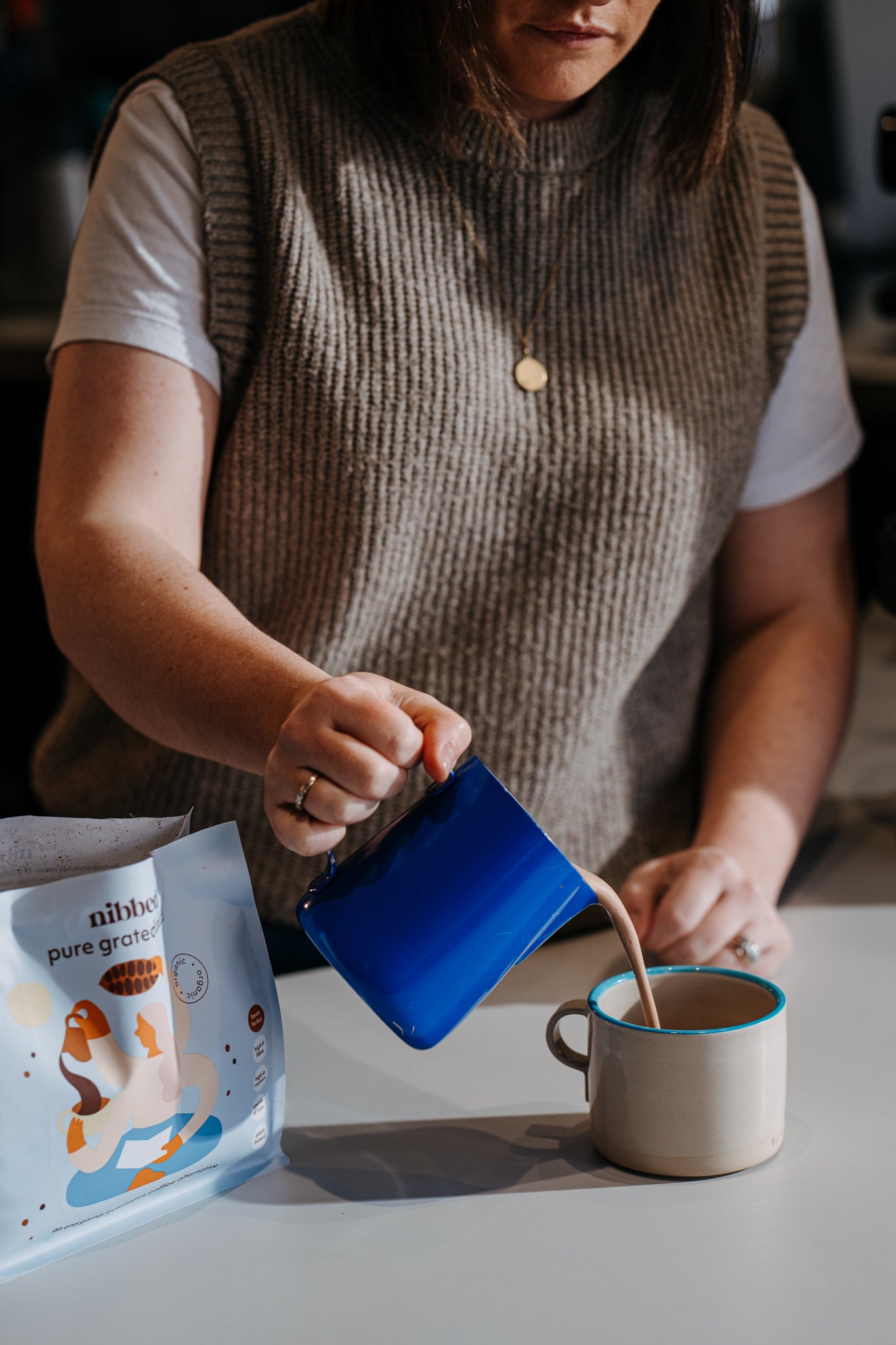Person pouring a blue mug into a white mug with a package of 'nibbed' pure cacao on table.