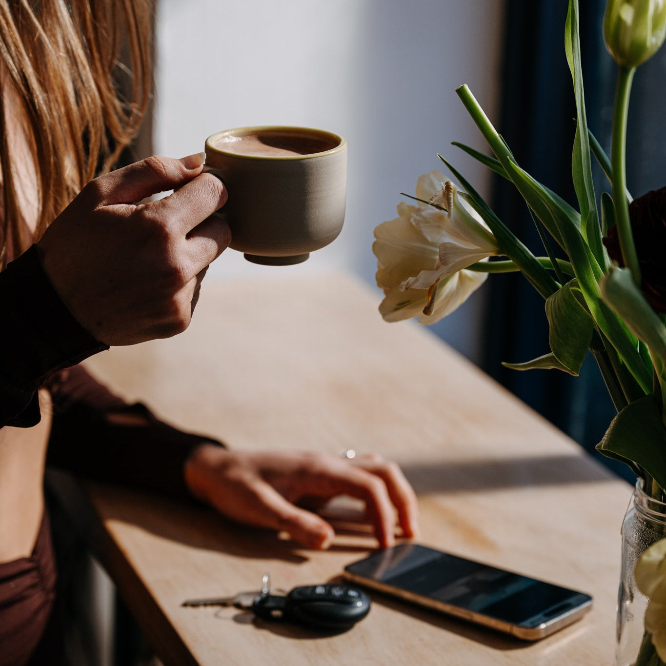 Person holding a nibbed cacao mug with a phone and keys on a table next to a vase with flowers.